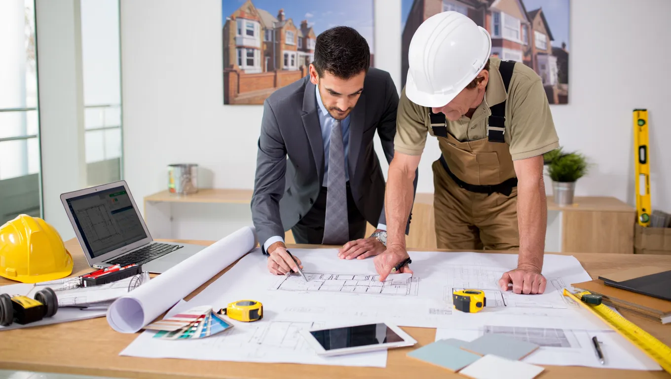 Property consultant and contractor reviewing HMO conversion blueprints with construction tools and laptop